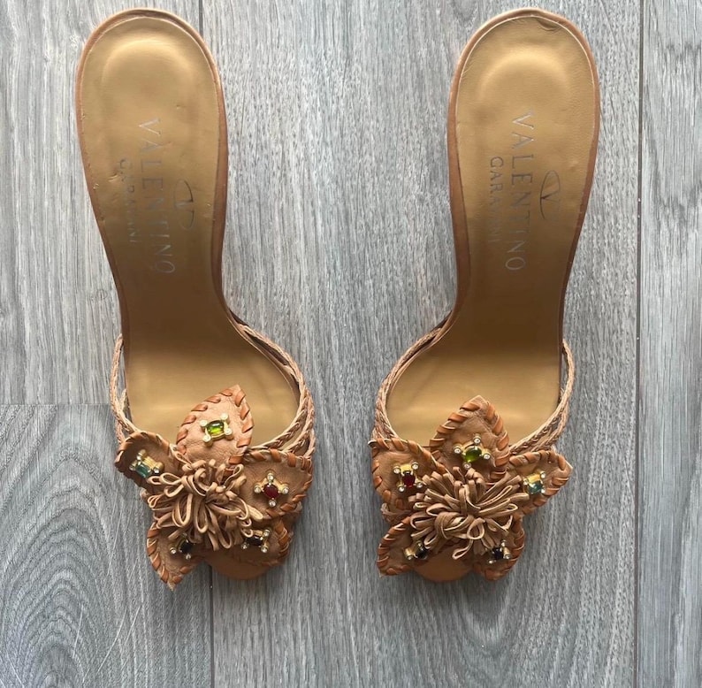 May include: A pair of tan leather Valentino Garavani heeled sandals. Each sandal features a floral embellishment with colourful jewel accents. The sandals have a braided strap design and a high heel. The Valentino Garavani logo is visible on the insole.