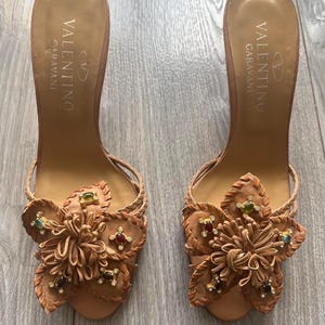 May include: A pair of Valentino Garavani brown leather heeled sandals. Each sandal features a decorative flower design with colourful jewel accents. The sandals have a braided ankle strap and the Valentino Garavani logo is printed on the insole.