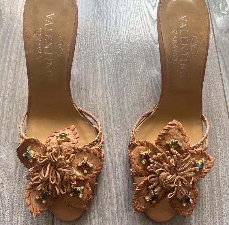 May include: A pair of Valentino Garavani brown leather heeled sandals. Each sandal features a decorative flower design with colourful jewel accents. The sandals have a braided ankle strap and the Valentino Garavani logo is printed on the insole.