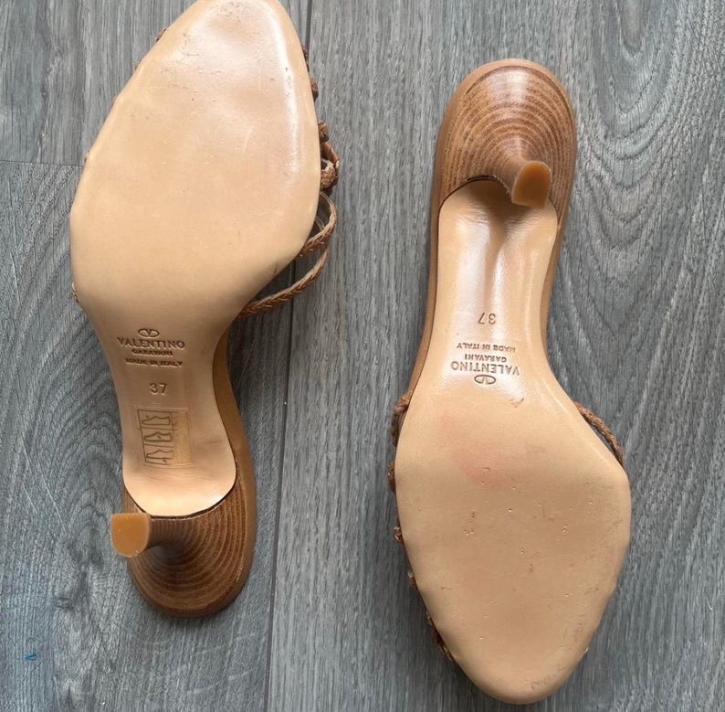 May include: Two brown leather heeled sandals with woven straps. The soles are light tan with the brand name "Valentino Garavani" and size 37. The heels are approximately 5 cm high.