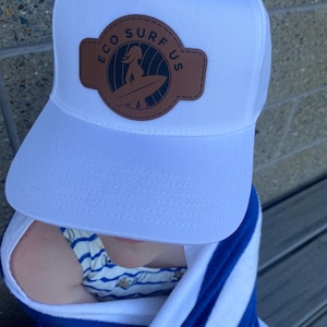 Op de afbeelding: Witte baseballcap met een bruin leren patch met de tekst "ECO SURF US" en een surferafbeelding. De cap heeft een gebogen klep en een mesh achterkant.