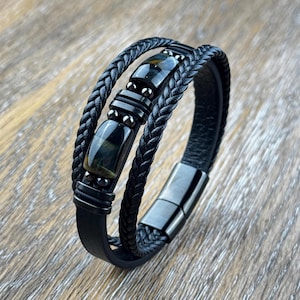 May include: Black leather bracelet with braided and smooth bands. The bracelet features rectangular and spherical dark blue tiger eye stones and a magnetic clasp. The bracelet is displayed on a wooden surface.