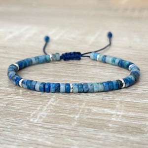 May include: A blue beaded bracelet with a sliding knot closure. The bracelet features small, square-shaped beads in shades of blue and silver accents. The bracelet is displayed on a light-colored wooden surface.