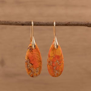 May include: Two teardrop earrings with gold-coloured hooks. The earrings have orange and yellow stones with a marbled pattern. They are suspended from a thin, brown branch against a blurred wooden backdrop.