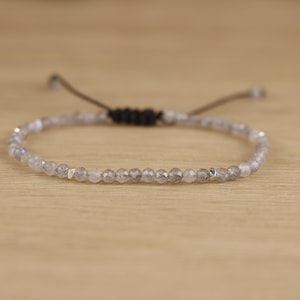 Op de afbeelding: Een delicate armband met kleine, facetgeslepen, grijze kralen geregen op een donker koord. De verstelbare armband heeft een zwarte geweven knoop en een schuifsluiting. De kralen zijn gelijkmatig verdeeld, waardoor een minimalistisch en elegant ontwerp ontstaat.