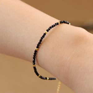 May include: A delicate bracelet made with black beads and gold accents. The bracelet is on a person's wrist.