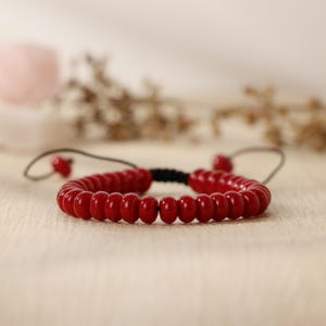 May include: A red beaded bracelet with a black cord and adjustable closure. The beads are small and round, and the bracelet is simple and elegant.