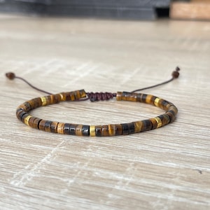 Könnte beinhalten: Armband mit kleinen, zylindrischen Perlen in Braun-, Gold- und Schwarztönen. Es hat einen verstellbaren Schiebeknotenverschluss. Die Perlen haben eine polierte Oberfläche. Das Armband ist für den täglichen Gebrauch geeignet.
