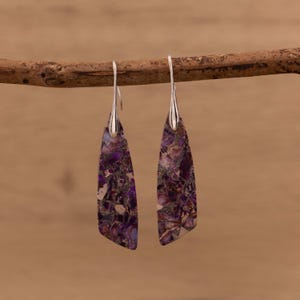May include: A pair of drop earrings featuring silver hooks and rectangular, purple stones with a marbled design. The earrings hang from a brown branch against a blurred backdrop.