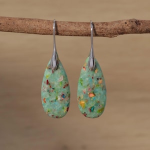 May include: Teardrop earrings with a speckled light green design. The earrings have silver-coloured hooks and are suspended from a brown branch. The speckled design includes red, yellow, and blue accents.