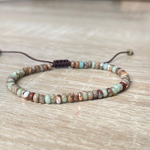 Könnte beinhalten: Ein Armband mit kleinen, runden Perlen in Braun-, Rot-, Grün- und Weißtönen. Die Perlen sind auf einem braunen Kordelband mit verstellbarem Verschluss aufgefädelt. Das Armband ist für das Handgelenk konzipiert.