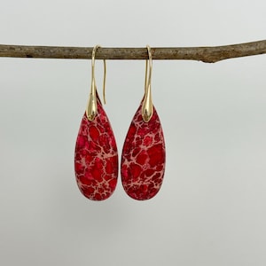 May include: A pair of teardrop-shaped earrings with a vibrant red and cream marbled pattern. The earrings have gold-colored ear wires and caps. The earrings are hanging from a brown branch against a white background.