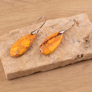 Puede incluir: Un par de pendientes en forma de lágrima con un vibrante patrón veteado amarillo y naranja. Los pendientes tienen ganchos plateados y curvados. Se exhiben sobre una superficie clara y texturizada de color marrón.