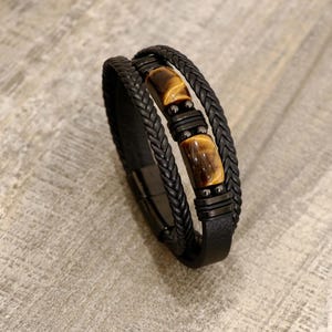 May include: Black leather bracelet with braided and smooth bands. The bracelet features two rectangular tiger's eye stones and black metal accents. The bracelet is designed with a magnetic clasp.