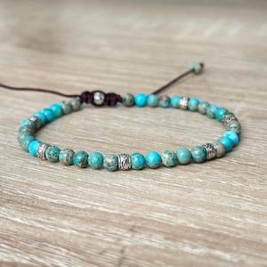 May include: A turquoise and grey beaded bracelet with silver-coloured accents. The bracelet is strung on a dark brown cord and features adjustable sizing. The beads have a mottled appearance, displayed on a light brown wooden surface.