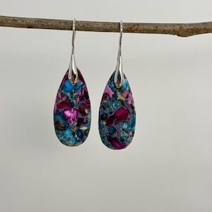May include: A pair of teardrop-shaped earrings with a mosaic pattern of blue, pink, and purple stones. The earrings are suspended from silver-colored hooks, creating a striking contrast with the colorful stones. The earrings are hanging from a small branch.