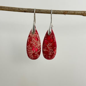 Op de afbeelding: Een paar druppelvormige oorbellen met een rood en beige gespikkeld patroon. De oorbellen hebben zilverkleurige haken. Ze hangen aan een bruine tak tegen een witte achtergrond.