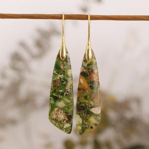 May include: Dangle earrings with gold-coloured tops and long, teardrop-shaped stones. The stones have a mottled pattern of green, brown, and beige. The earrings are suspended from a thin, brown rod.