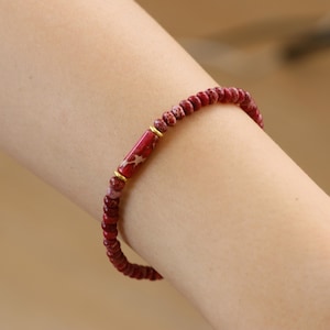 May include: A red and white beaded bracelet with a gold accent. The bracelet is made of small, round beads and is worn on a person's wrist.