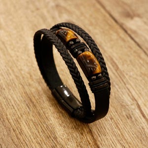 May include: Black leather bracelet with braided accents and tiger eye stone beads. The bracelet features a magnetic clasp and a multi-strand design. The tiger eye stones have a warm brown color.