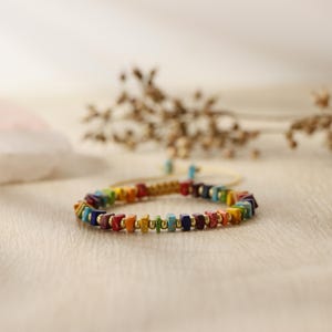 May include: A rainbow colored beaded bracelet with gold accents. The bracelet is made of small square beads and is strung on a gold chain.
