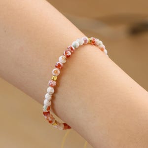 Puede incluir: Un brazalete de cuentas de ágata blanca y roja con detalles dorados. El brazalete está en la muñeca de una persona.