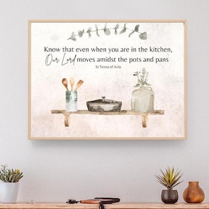 May include: A watercolor print with a quote about faith in the kitchen. The quote reads "Know that even when you are in the kitchen, Our Lord moves amidst the pots and pans." The quote is attributed to St. Teresa of Avila. The print features a watercolor illustration of a kitchen shelf with a pot, utensils, and a vase of flowers.