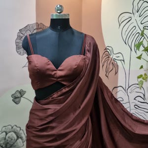 May include: A dark brown satin saree and matching blouse displayed on a black mannequin. The blouse has a sweetheart neckline and spaghetti straps. The saree is draped to show the fabric's flow and texture. The background features a wall with floral and leaf designs.