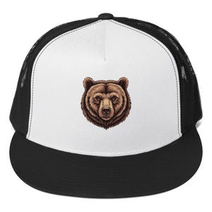 May include: A black and white trucker hat with a detailed brown bear face graphic on the front. The hat features a white front panel and a black mesh back, with a curved black brim. The bear graphic is centered on the front panel.