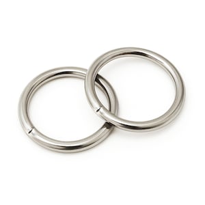 Op de afbeelding: Twee cirkelvormige metalen ringen met een gladde, gepolijste zilverkleurige afwerking. Elke ring heeft een zichtbare naad waar de uiteinden samenkomen, wat zorgt voor een doorlopend, afgerond profiel. De ringen zijn in elkaar gehaakt en overlappen elkaar.