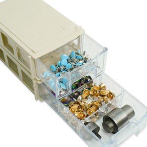 May include: A beige plastic storage unit with clear drawers, filled with various craft supplies. The drawers contain light blue embellishments, iridescent beads, and gold-coloured metal fasteners. A metal tool is also visible.