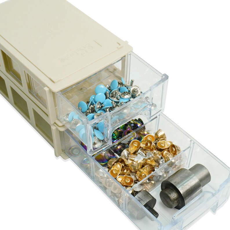 May include: A beige plastic storage unit with clear drawers, filled with various craft supplies. The drawers contain light blue embellishments, iridescent beads, and gold-coloured metal fasteners. A metal tool is also visible.