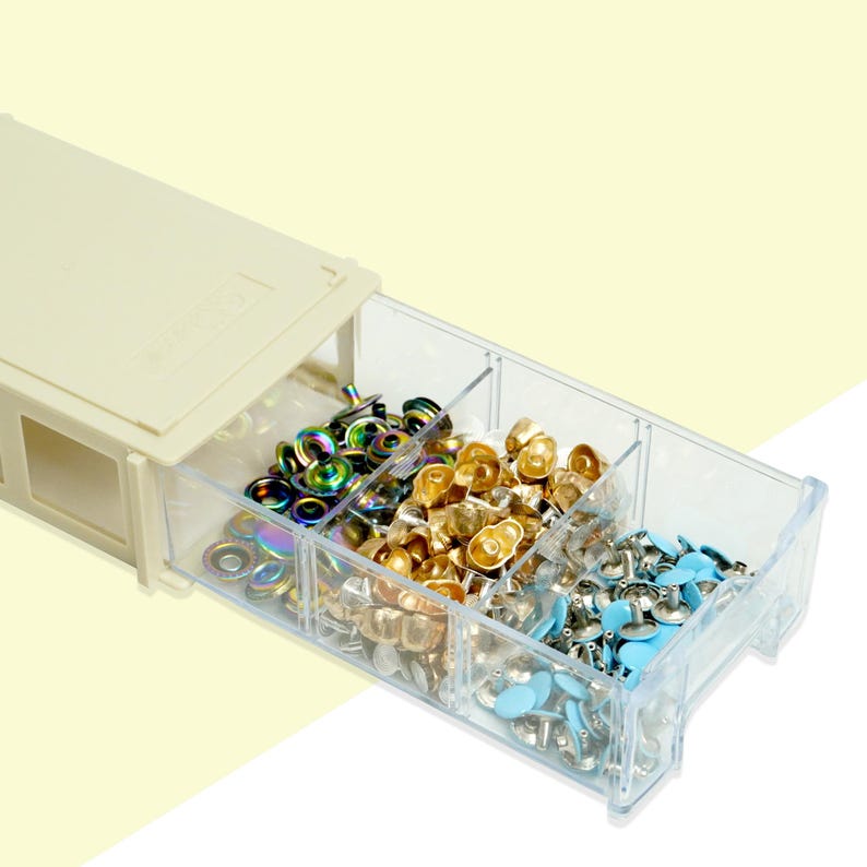 May include: A beige plastic organiser with a clear, divided drawer filled with colourful metal snaps. The snaps are in iridescent, gold, silver, and light blue colours, ideal for craft projects and sewing.