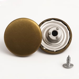 May include: Two round metal buttons with a matte antique brass finish. One button shows the smooth, solid front surface, while the other displays the silver-toned metal underside with a central attachment mechanism. A small silver screw is included.