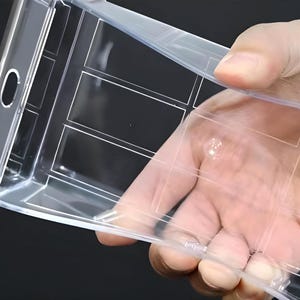 May include: A clear plastic container, rectangular in shape, with a grid pattern. The container is held in a hand and has a smooth, transparent surface. It features a curved opening on the side. The container appears empty.