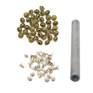 May include: A collection of brass-toned conical studs and silver-toned metal rivets arranged in two separate groups, accompanied by a single cylindrical metal setting tool used for the application of these hardware fasteners.