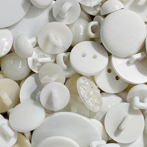 May include: A collection of assorted white buttons in various sizes and shapes. The assortment includes round buttons with two holes, shank buttons with loops on the back, and textured surfaces. The buttons are opaque, glossy, and matte white.