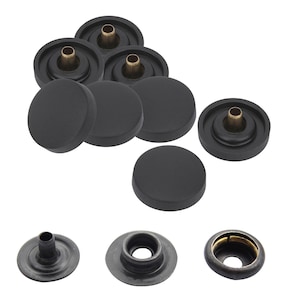 Può includere: Una collezione di bottoni a pressione in metallo nero con calotte circolari e perni color ottone sul retro. Il set include diversi componenti assemblati e singoli pezzi metallici con fori centrali, il tutto presentato su uno sfondo bianco.