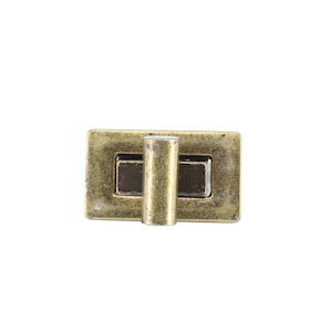 May include: A rectangular metal twist lock closure with a distressed antique brass finish. The hardware features a central rotating cylindrical knob mounted on a flat rectangular base plate, designed for securing bags or small cases with a turn mechanism.