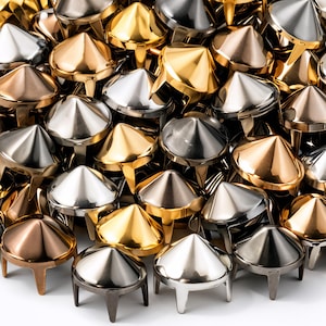 Può includere: Primo piano di una collezione di borchie coniche metalliche in varie finiture metalliche, tra cui oro, argento e bronzo. Le borchie hanno una base a tre punte e una parte superiore appuntita. L'immagine è ben illuminata, mostrando le superfici riflettenti.