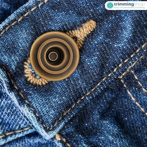 Può includere: Primo piano di un bottone color bronzo su tessuto denim blu. Il bottone è circolare con anelli concentrici e un foro centrale. Le cuciture beige accentuano il bottone e le cuciture del denim.