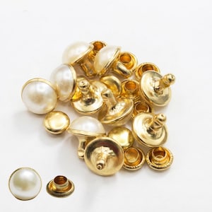 May include: A collection of gold-toned metal buttons featuring a smooth, round, off-white faux pearl top. Each button has a metallic base with a central post for attachment. The set includes multiple individual pieces shown from various angles.