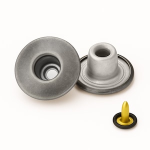 Könnte beinhalten: Zwei kreisförmige Metalldruckknopf-Komponenten mit mattgrauem Finish und einer zentralen hohlen Öffnung. Daneben befindet sich ein kleineres, separates Befestigungsteil mit einer goldfarbenen Spitze und einem schwarzen Ring.