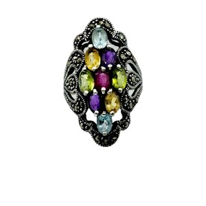 May include: A silver-toned ring with an oval design, featuring a cluster of colorful gemstones. The gemstones include blue, yellow, purple, and green hues. The ring has a detailed, ornate setting with small, dark accents.
