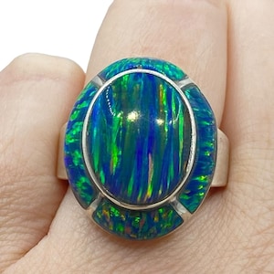 May include: A silver ring featuring a large, oval-shaped opal. The opal displays vibrant blue and green hues with a striking, iridescent pattern. The ring's setting is silver, enhancing the gemstone's colors.