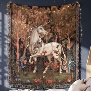 May include: A woven wall tapestry featuring a white unicorn standing in a dense forest with dark foliage and red flowers. The rectangular textile has a fringed edge and displays a detailed scene of the mythical creature amidst trees and plants.