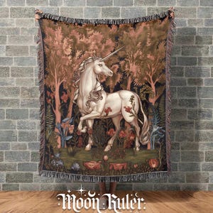 May include: A woven throw blanket featuring a white unicorn standing in a forest with pink and brown trees, green foliage, and red flowers. The rectangular blanket has a fringed edge and a detailed tapestry-style design with a dark background.