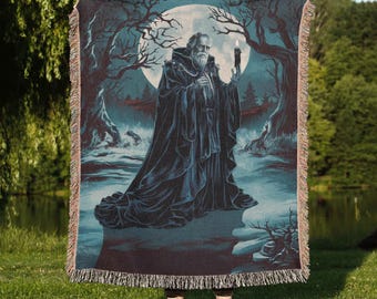Op de afbeelding: Een donkerblauwe en blauwgroene geweven sprei met een gedetailleerde illustratie van een oude man die een kaars vasthoudt in een bosrijke omgeving met een volle maan. De sprei heeft een gefranjerde rand en de woorden "Moon Ruler" staan onderaan.