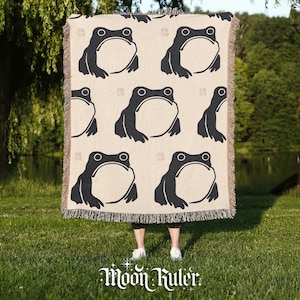 May include: A woven throw blanket featuring a repeating pattern of dark charcoal frogs on a light beige background. The blanket has a textured, fringed edge on the top and bottom, and the design includes small, subtle square icons between the frogs.