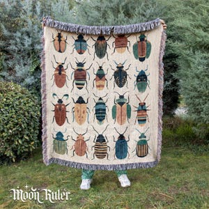 May include: Beige throw blanket with a repeating pattern of colourful beetles. The blanket has a fringed edge and is held up against green foliage. The Moon Ruler logo is visible at the bottom.
